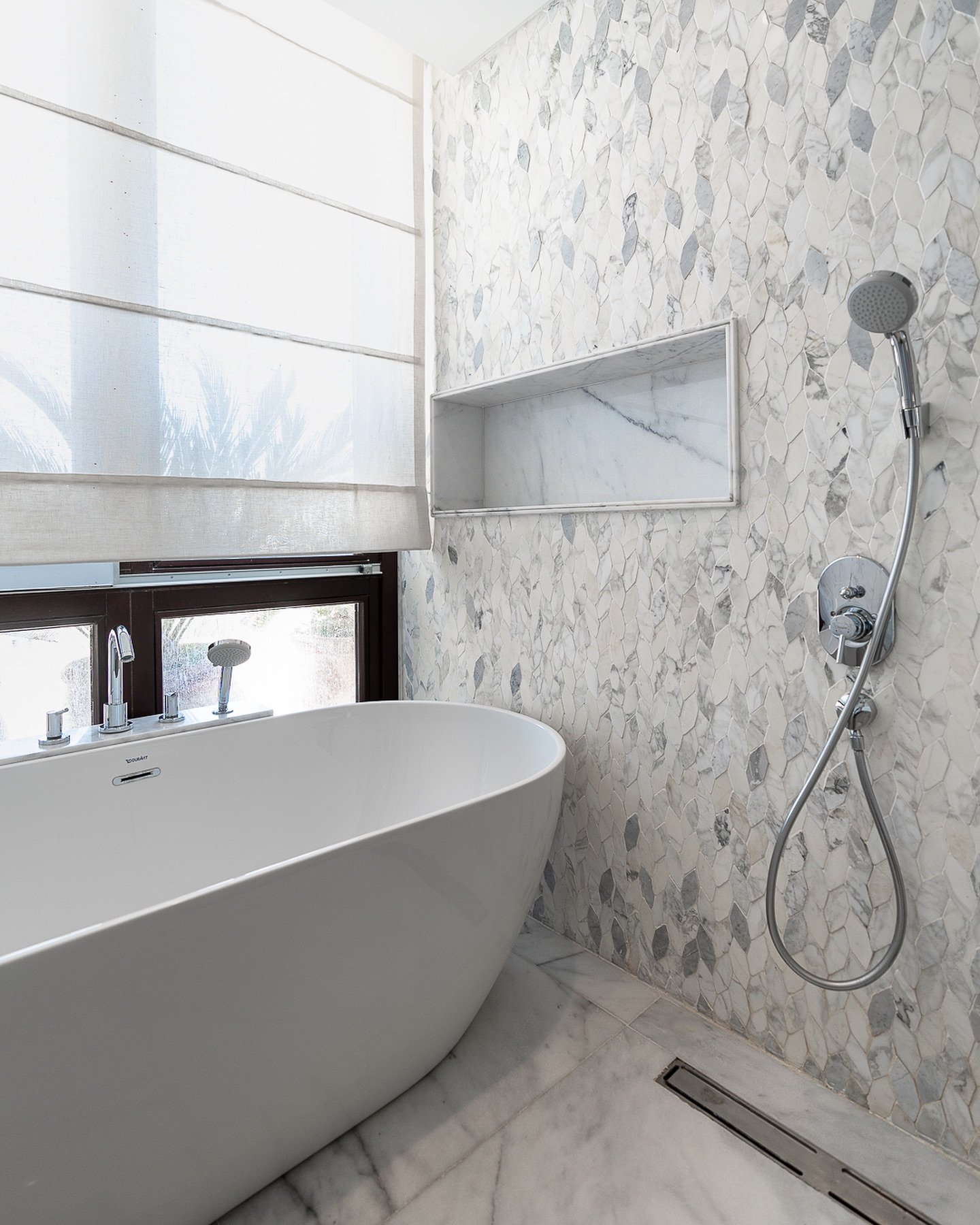 White freestanding bathtub against a wall of small, irregular marble mosaic tiles with a niche.