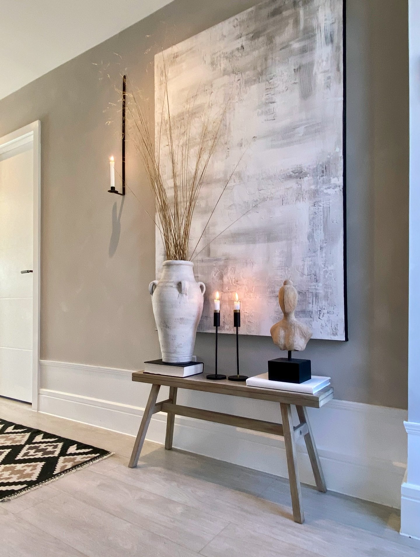 A hallway features a large white abstract painting above a rustic wooden bench with a tall vase and candles against a beige wall.