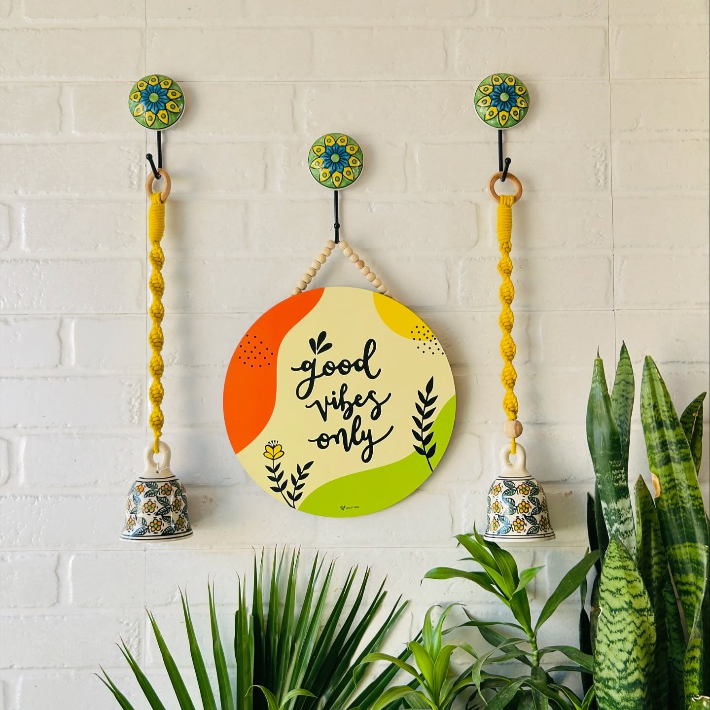 A white painted brick wall features a round "good vibes only" sign, ceramic floral hooks with yellow hanging bells, and snake plants.