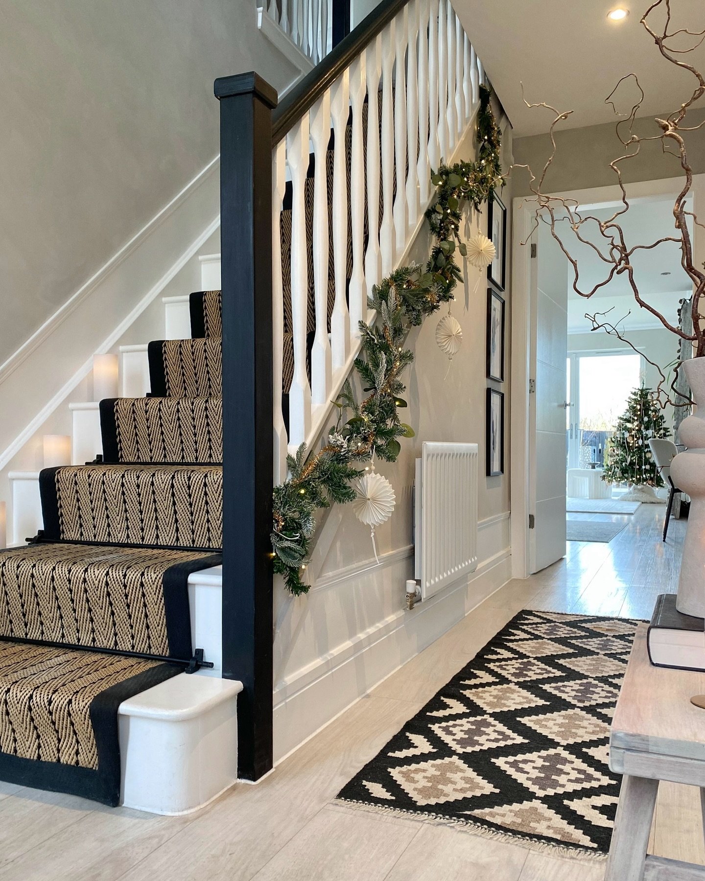 A hallway staircase wall features black framed art, white wainscoting, and a festive garland wrapped around the banister.