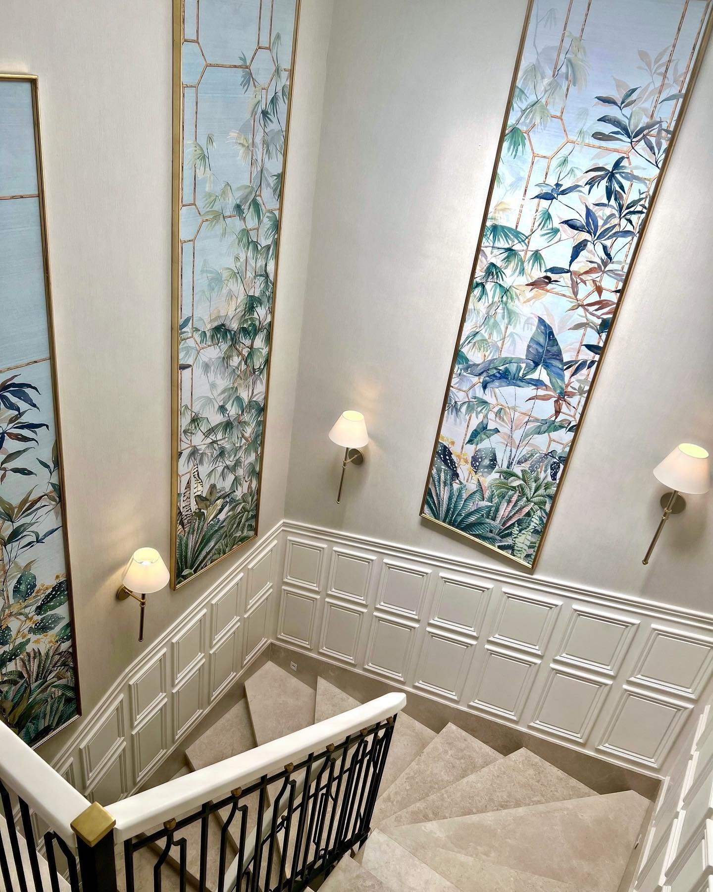 Staircase wall featuring white wainscoting and large framed tropical botanical art panels with brass sconces.