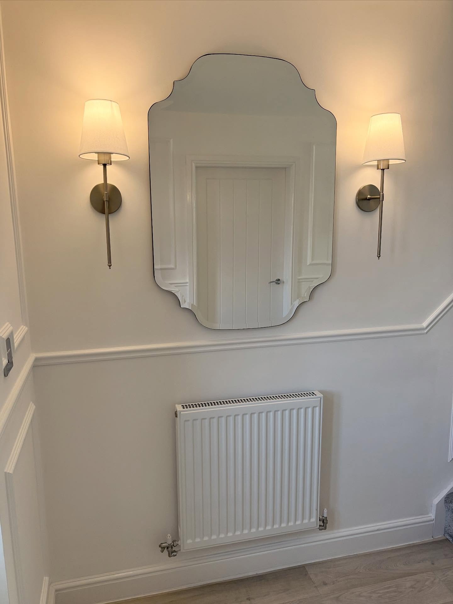 A white hallway wall features a shapely frameless mirror flanked by silver wall sconces above a white radiator.