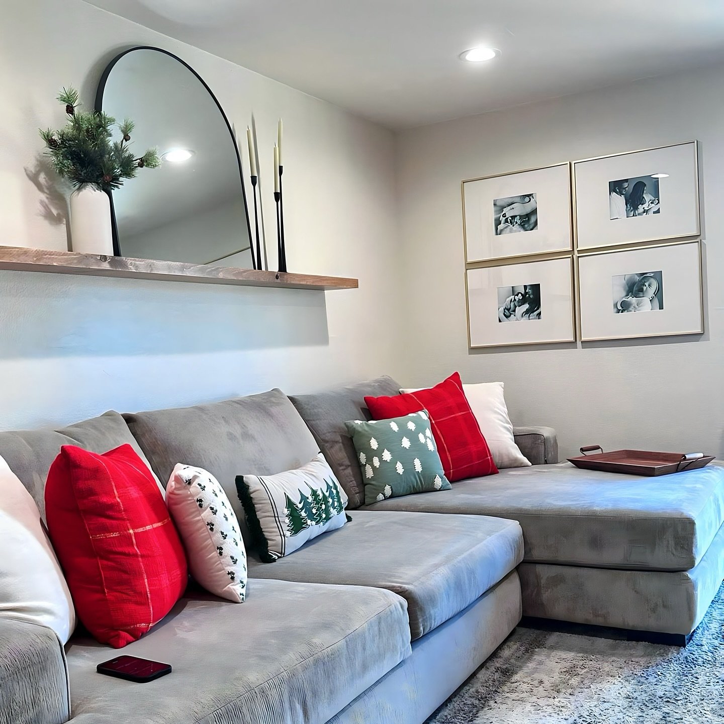 Floating wood shelf with mirror next to grid of four gold frames above grey sectional.