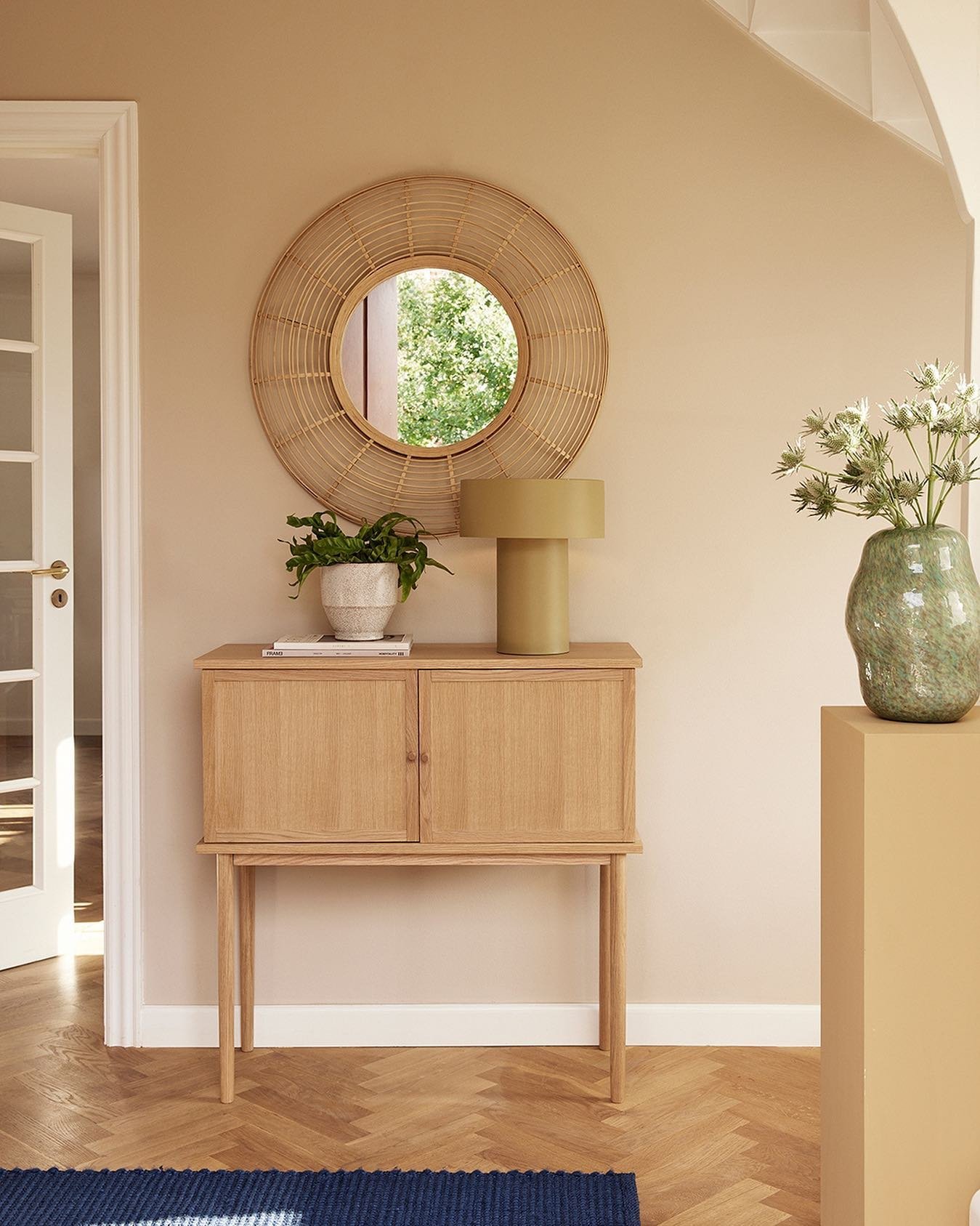 A beige hallway wall features a round rattan mirror hung above a light oak console table with a green lamp and vase.