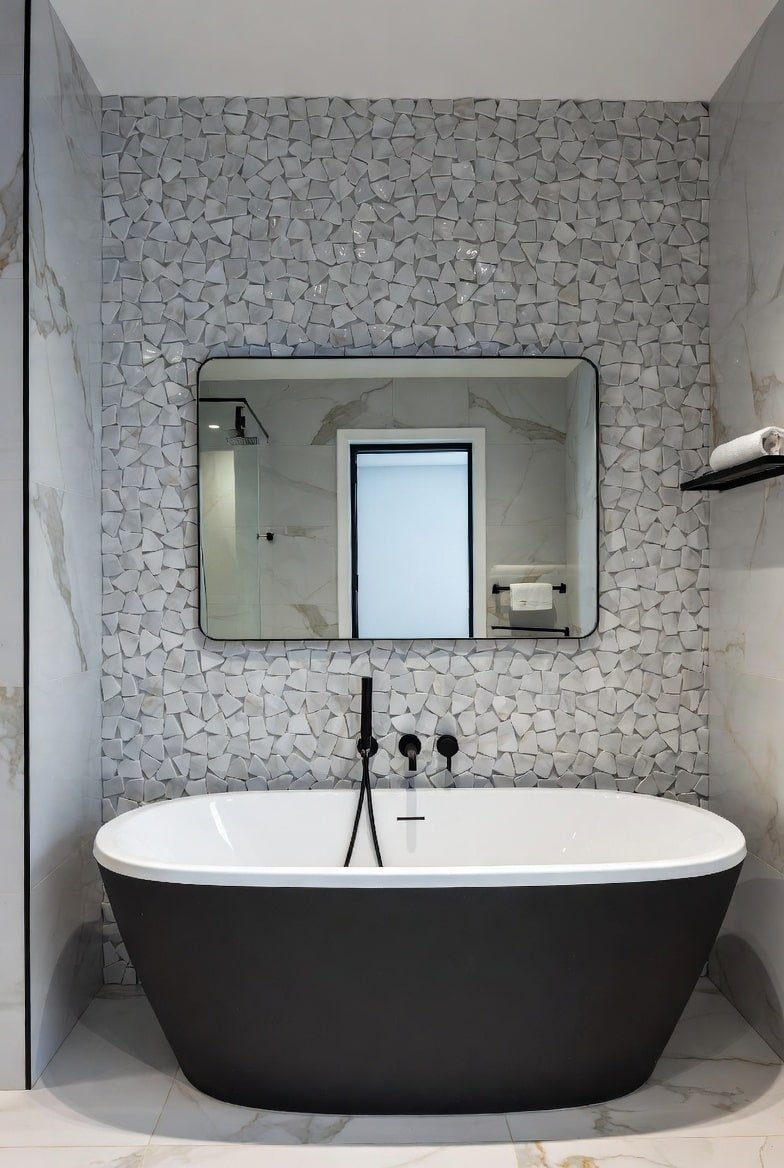 Black freestanding tub placed in front of a white wall featuring irregular broken mosaic tiles.