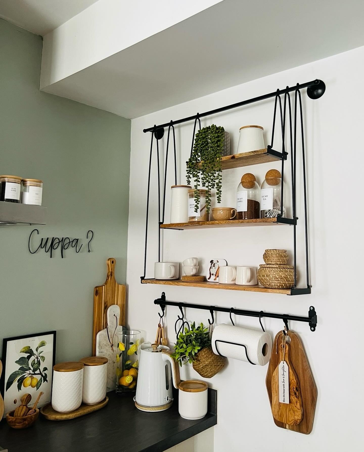 White kitchen wall featuring black industrial hanging shelves next to a sage green accent wall with wire text