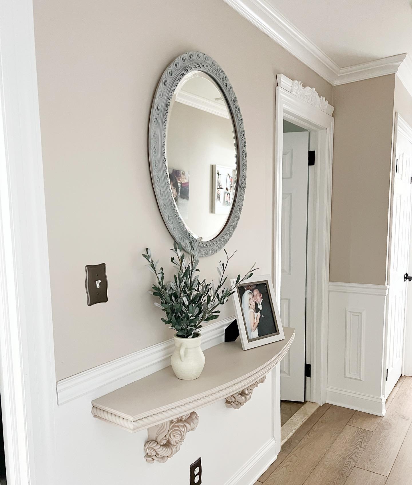 A beige hallway wall features an ornate oval mirror hanging above a curved floating shelf supported by decorative corbels with greenery.