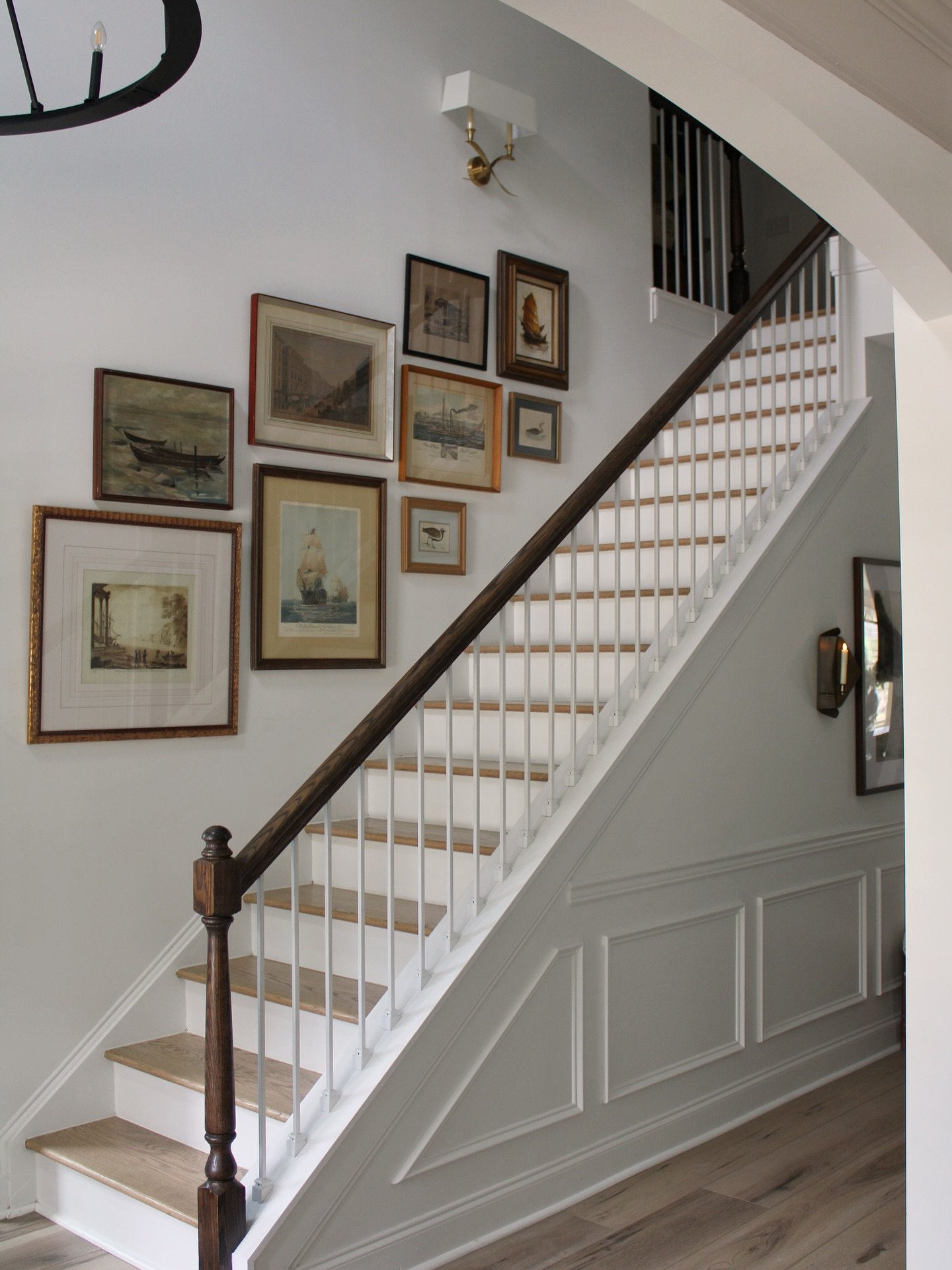 White staircase wall decorated with an eclectic gallery of vintage framed art above white wainscoting panels.