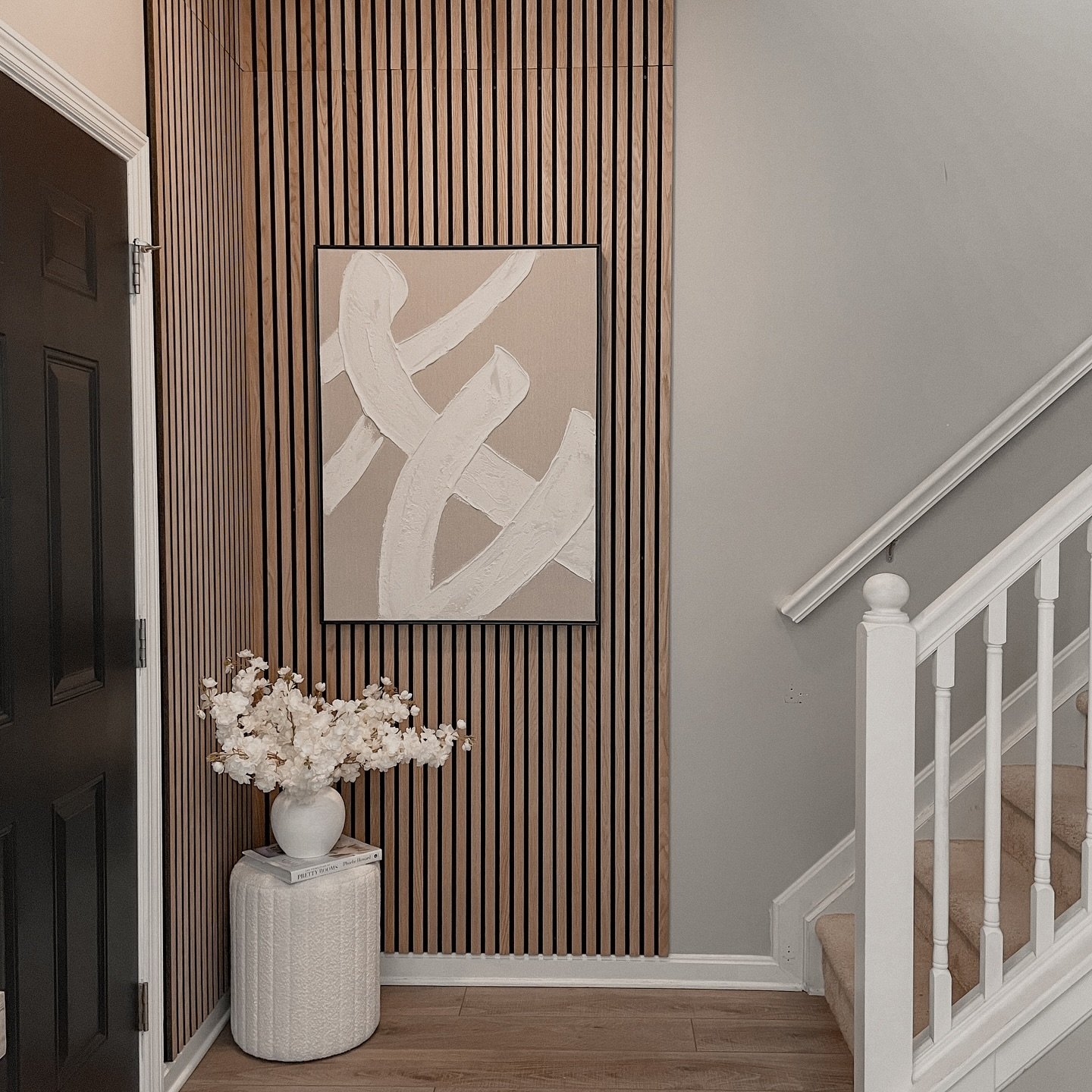 Entryway corner featuring vertical wood slats, beige abstract painting, and white textured stool with flowers near staircase.