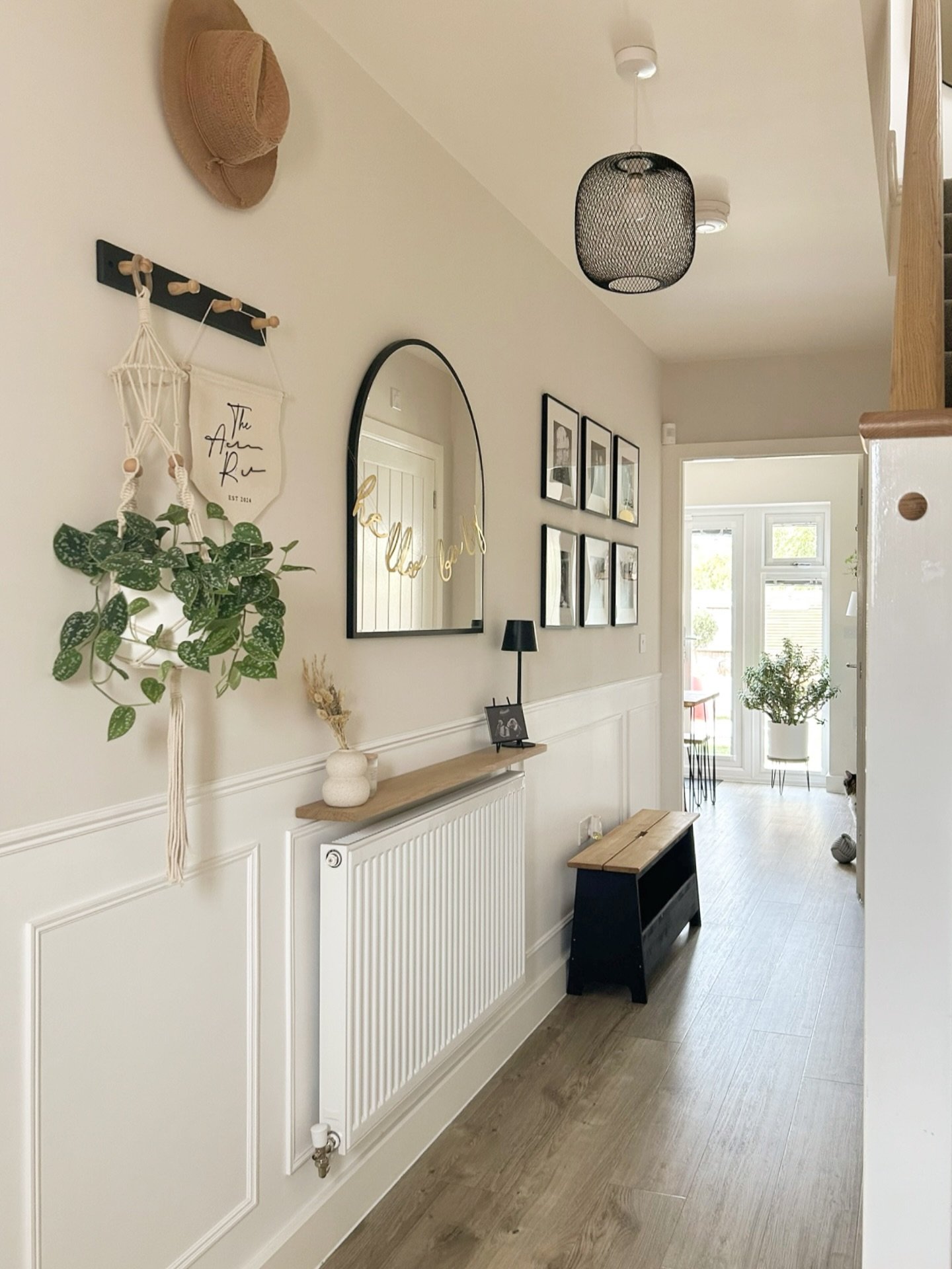 A narrow hallway features white wainscoting, an arched mirror, hanging plants, and black framed photos on a beige wall.