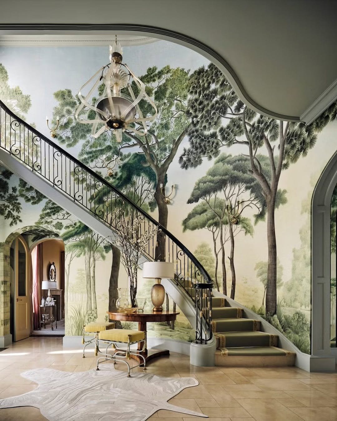 Grand curved staircase featuring a floor-to-ceiling scenic wallpaper mural of tall green trees and a landscape with brass sconces.