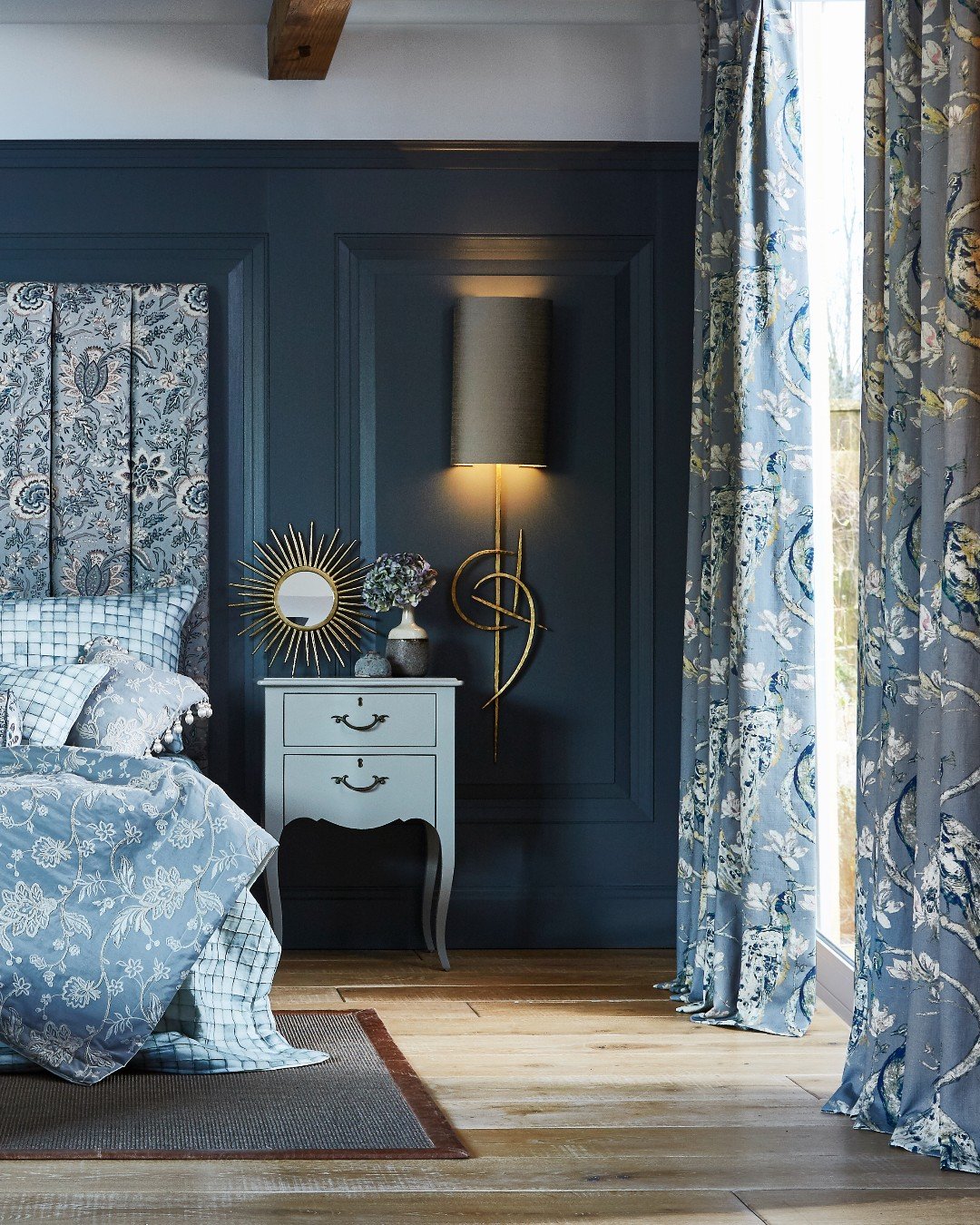 Navy blue paneled wall with gold sconce and floral curtains behind bed