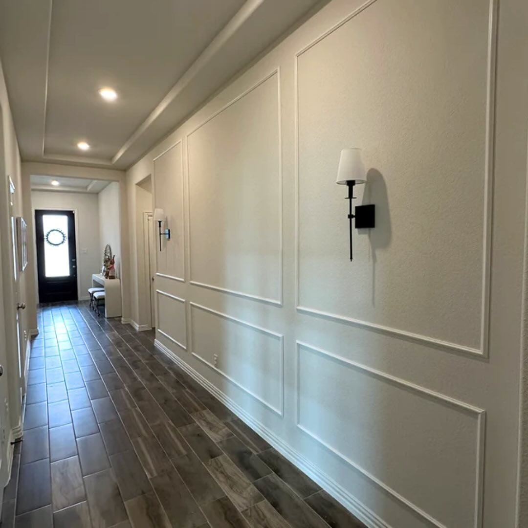 Long hallway accent wall featuring white picture frame molding, modern black sconces, and dark wood tile flooring.