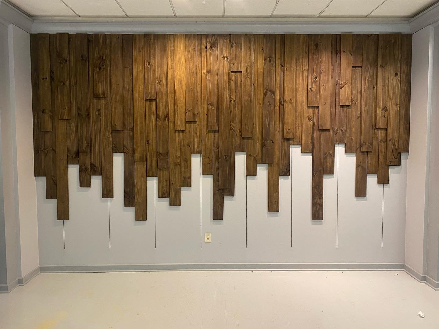 Vertical dark wood planks of varying lengths mounted on a white wall creating a dripping effect.