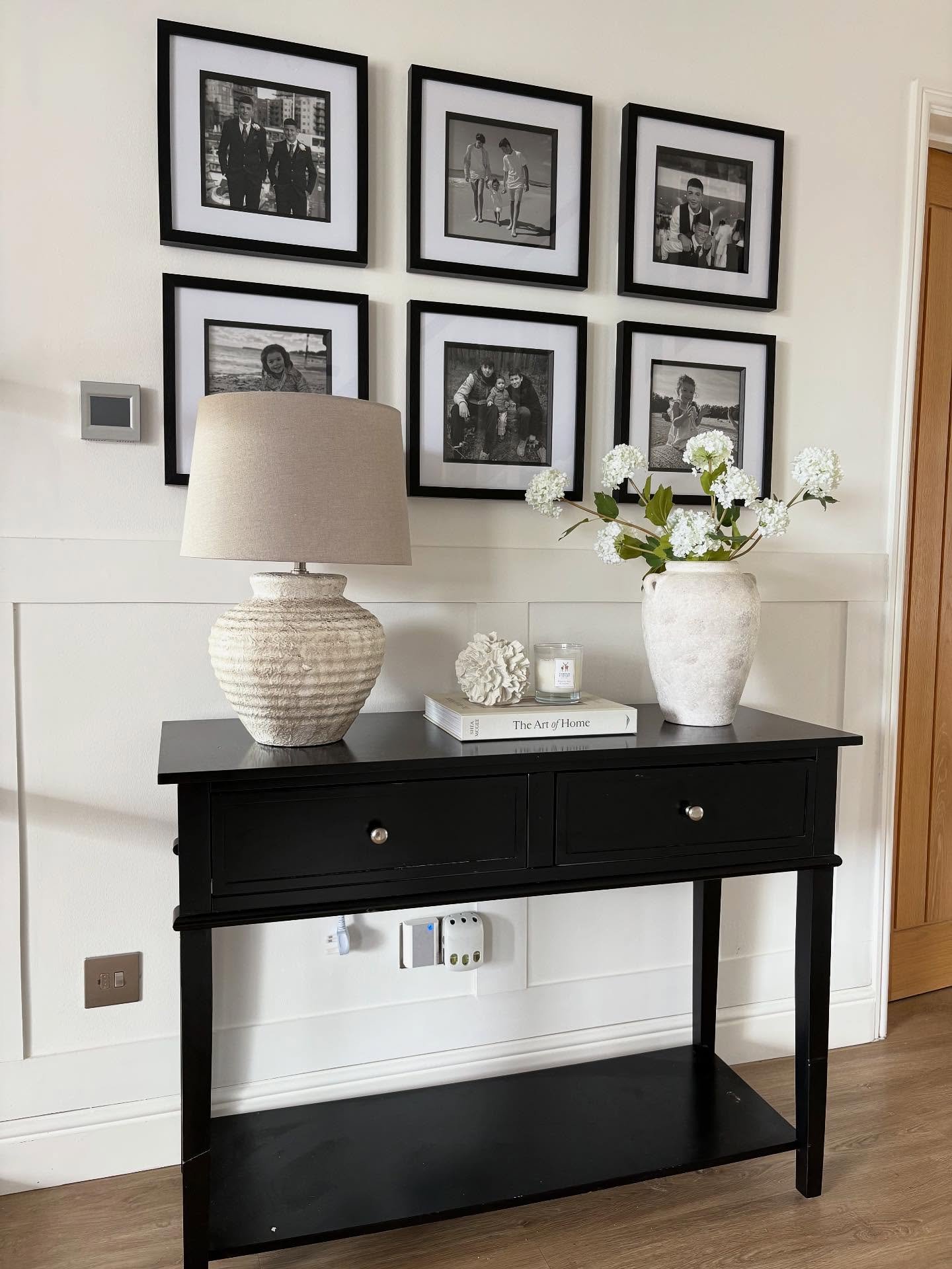 A hallway wall features a symmetrical grid of six black-and-white photos in black frames hung above a black console table with a textured lamp.