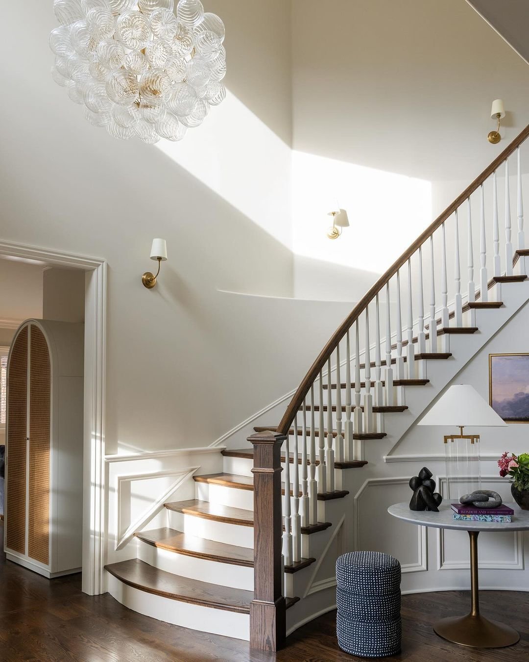 Bright white staircase featuring raised panel wainscoting, brass wall sconces with linen shades, and a large glass bubble chandelier.