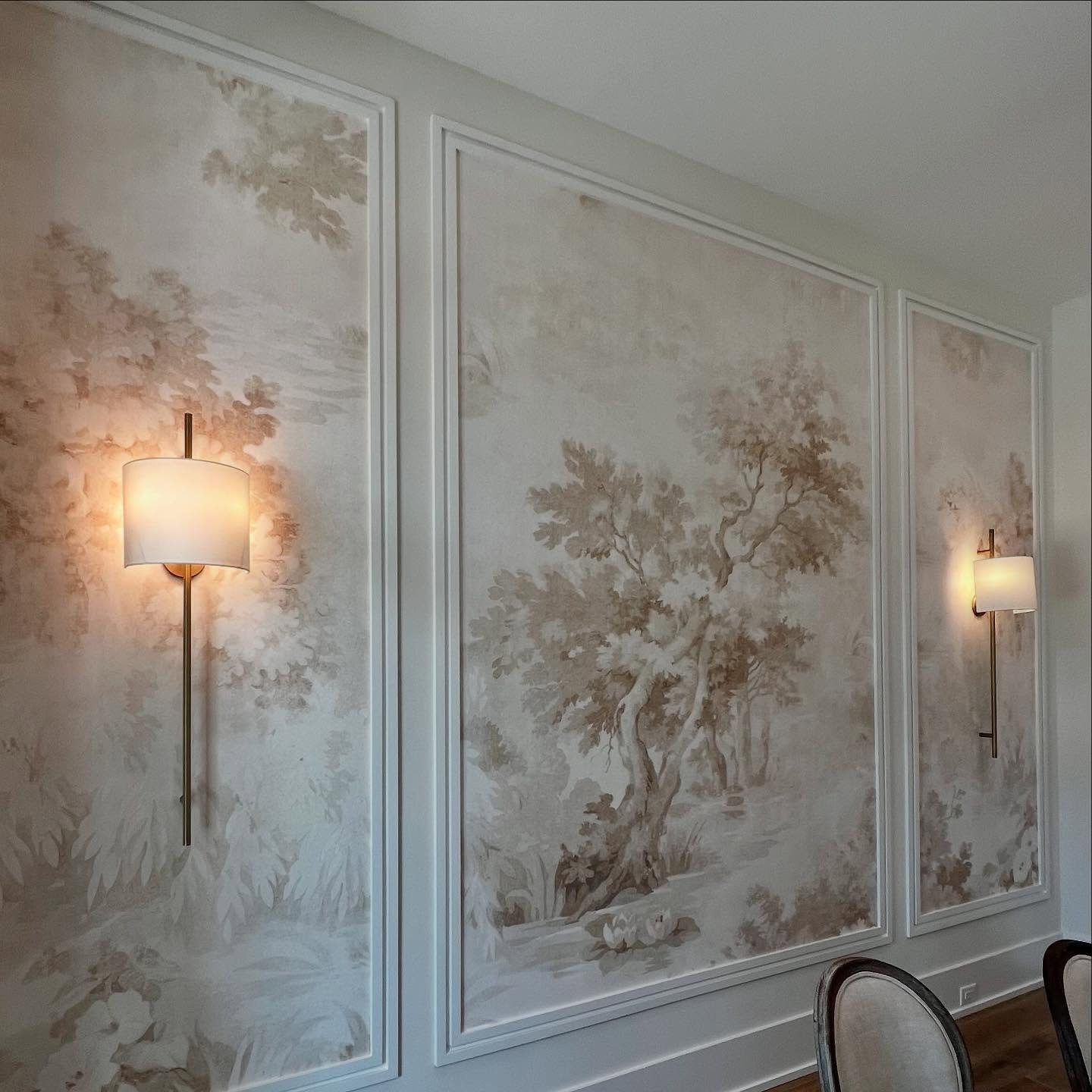 Dining room accent wall featuring sepia tree mural wallpaper framed by white molding with sconces.