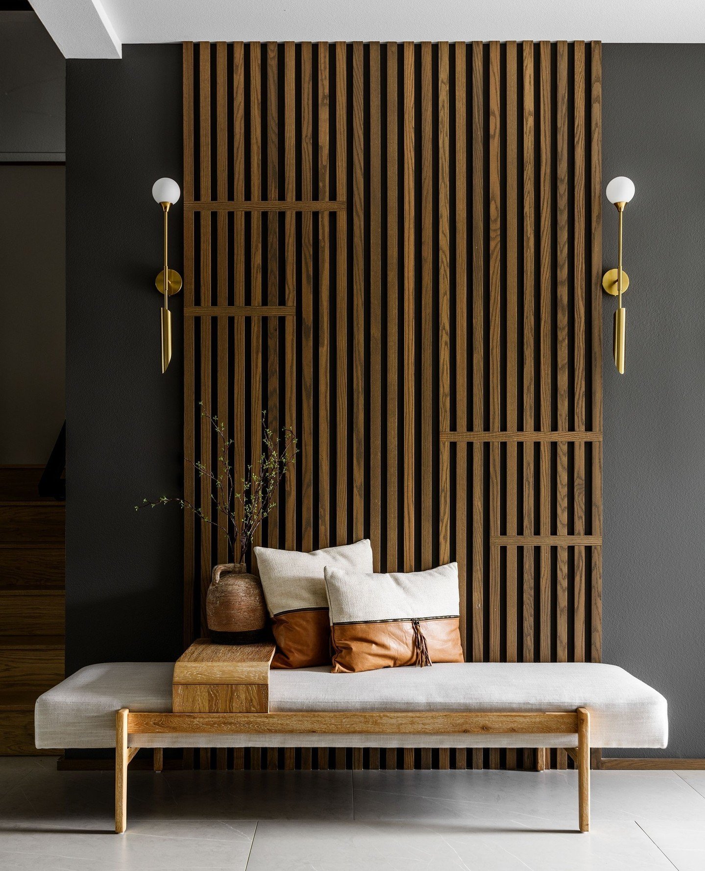Modern entryway with dark grey wall, geometric wood slat panel, gold sconces, and upholstered bench.