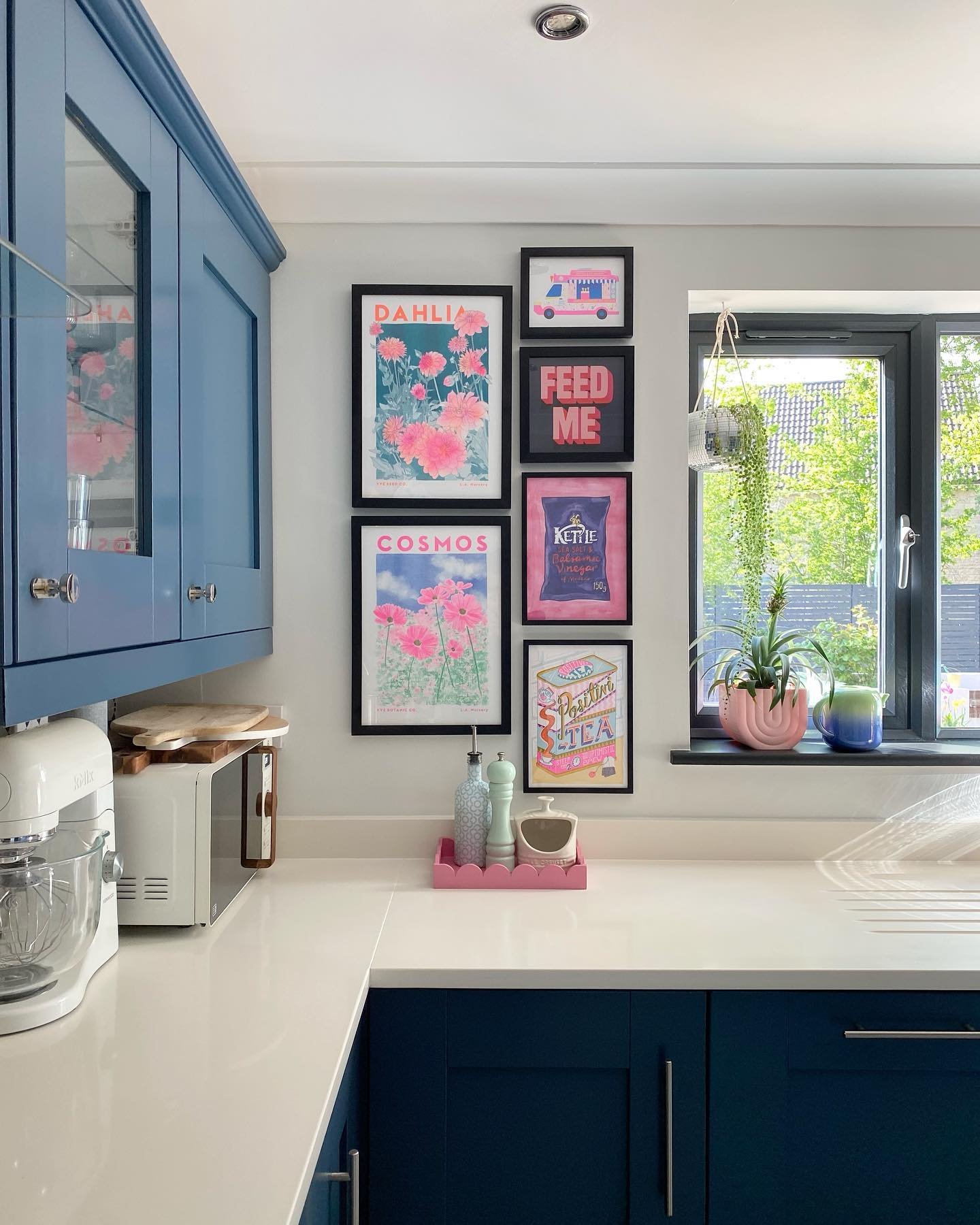 Kitchen corner featuring a gallery wall of pink floral and food-themed art prints next to blue cabinets