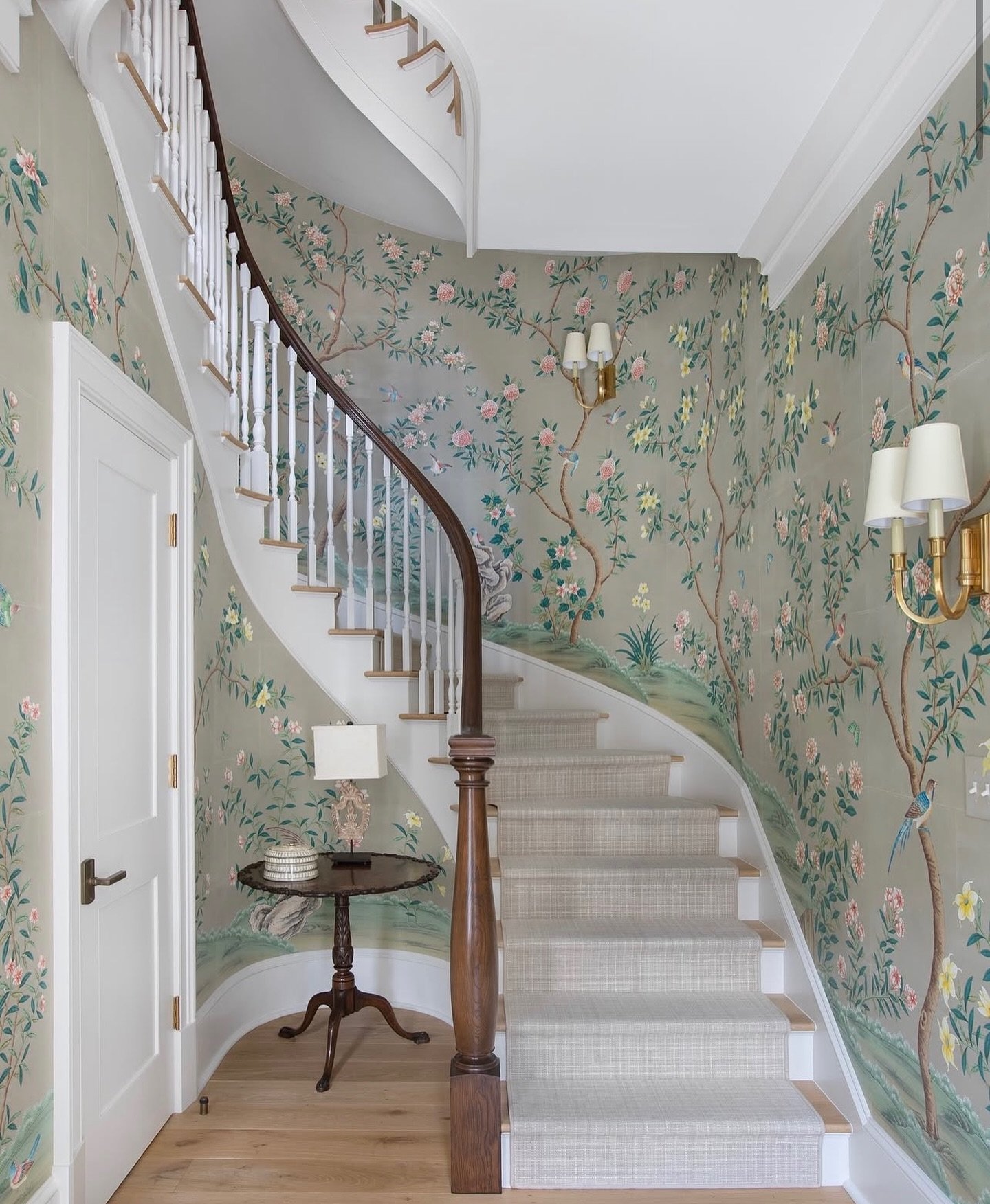 Curved staircase wall covered in sage green floral Chinoiserie wallpaper with birds and brass sconces.