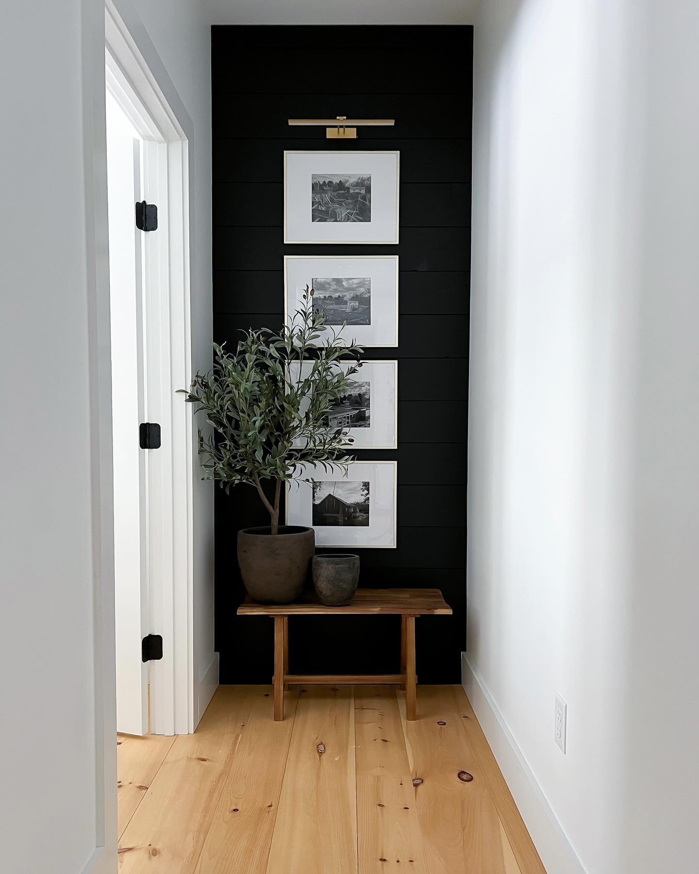 A narrow hallway end features a black horizontal shiplap wall with three stacked frames, a brass picture light, and a potted tree on a wooden bench.