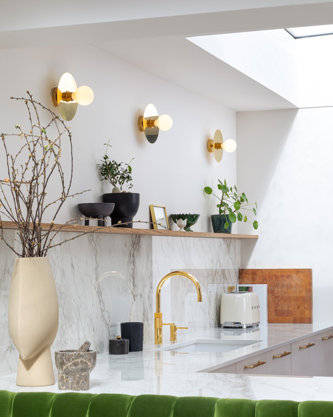 Three round gold wall lights mounted above a wooden kitchen shelf with marble backsplash.