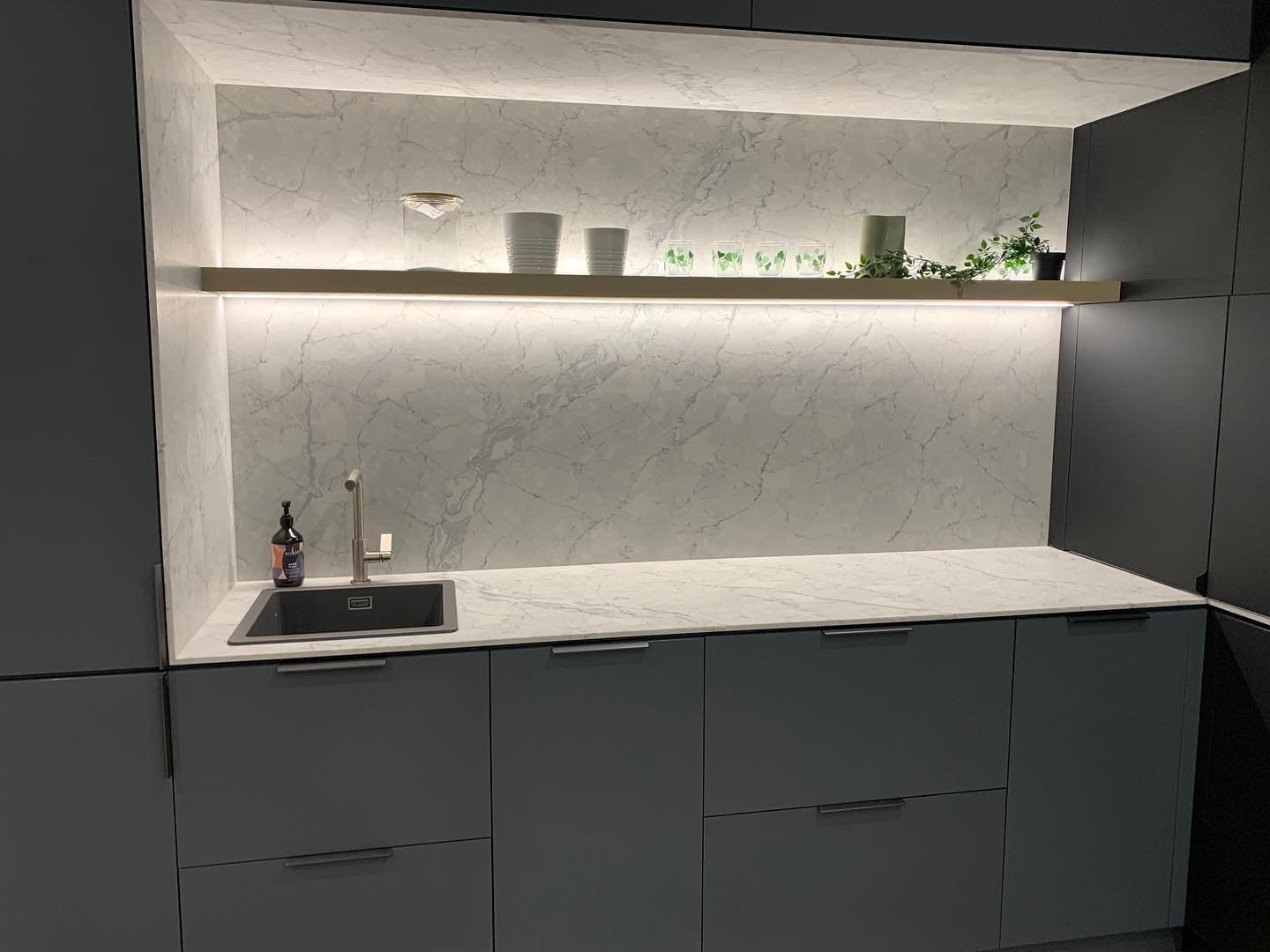 Dark grey kitchen cabinets with a white marble backsplash lit by an under-shelf LED strip over the sink.