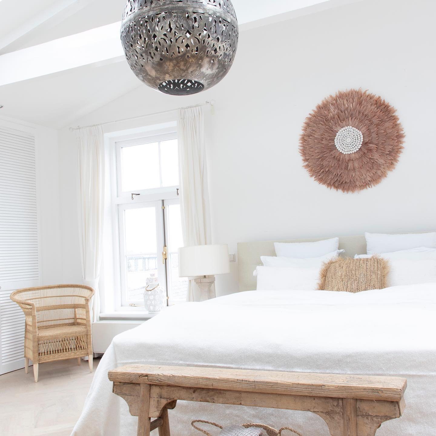 Large brown feather juju hat hanging above white bed in bright room