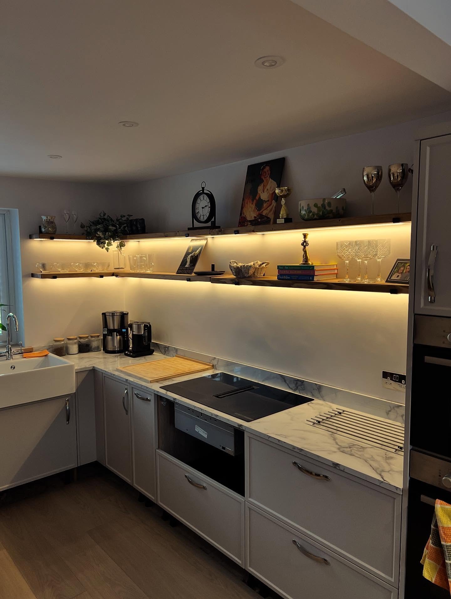 Kitchen corner featuring open wooden shelves with warm LED strips underneath, illuminating glassware and decor above white cabinets.