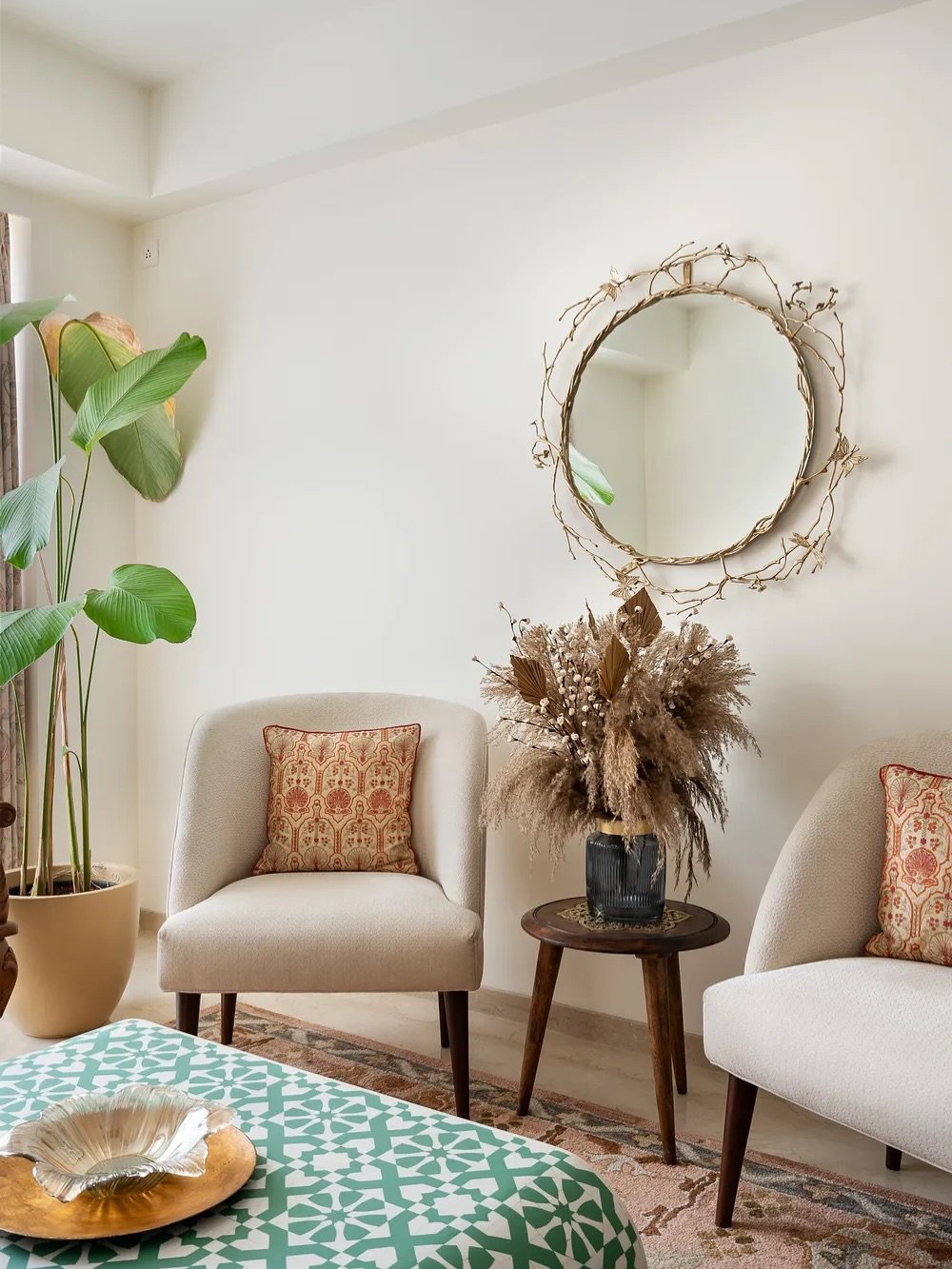 Round mirror with a gold branch-like frame hanging above two beige armchairs and a wooden side table with dried flowers.
