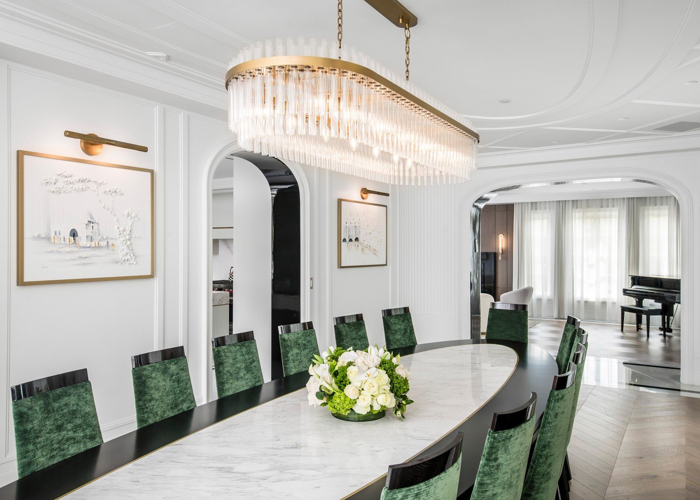 Long gold and crystal linear chandelier hanging over a marble dining table with green velvet chairs.