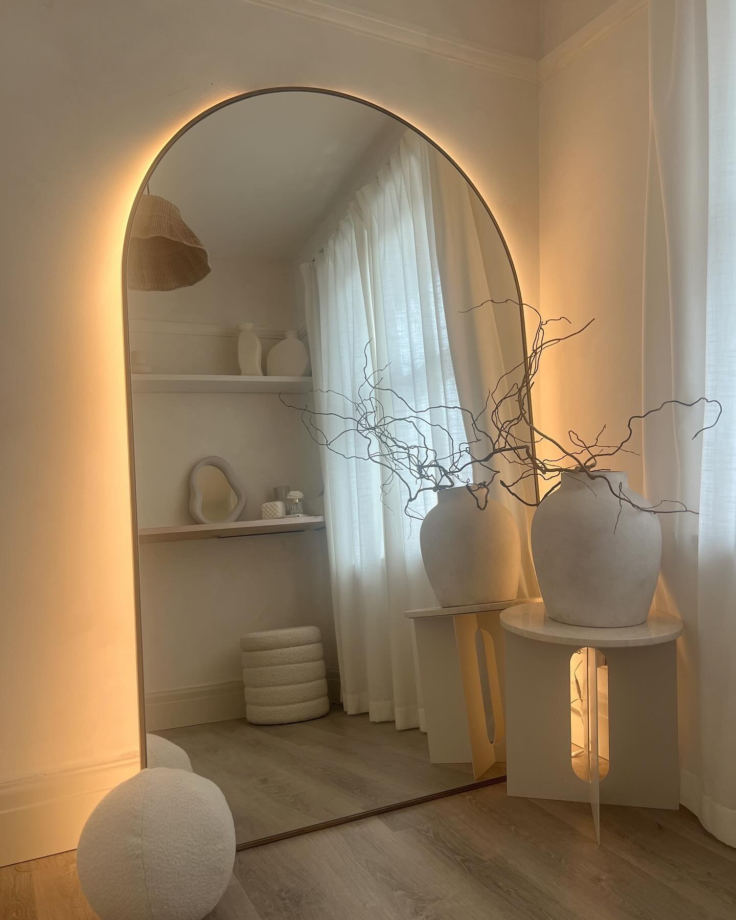 Large arched floor mirror leaning against a white wall with warm LED backlighting creating a soft halo effect.