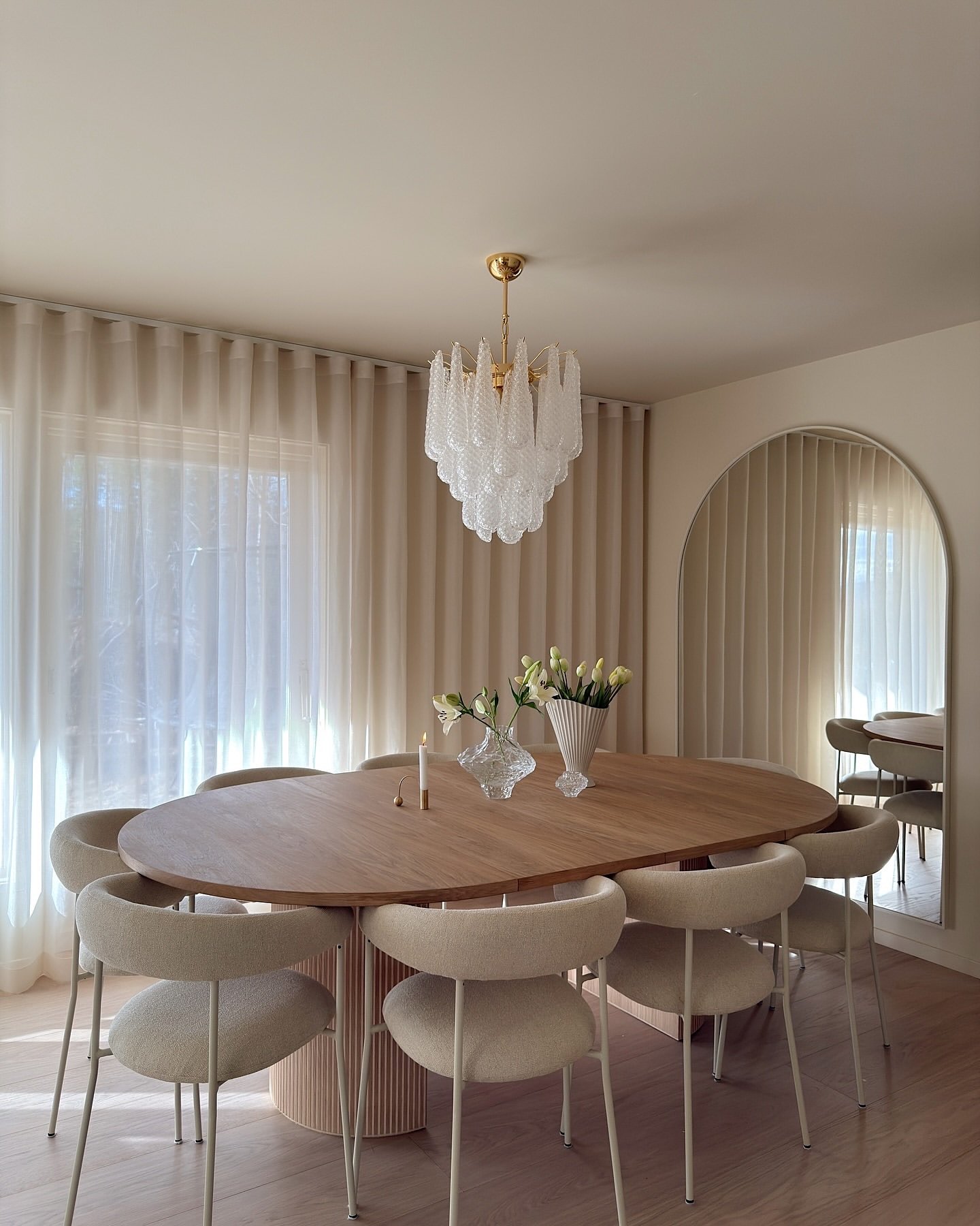 White textured glass chandelier over oval wood dining table with arched mirror and sheer curtains.