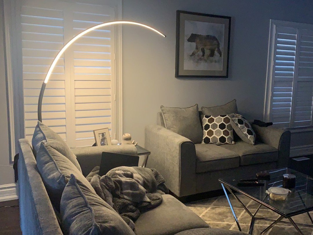 Cozy living room corner featuring a large LED arc floor lamp illuminating a grey wall with bear artwork and white window shutters.