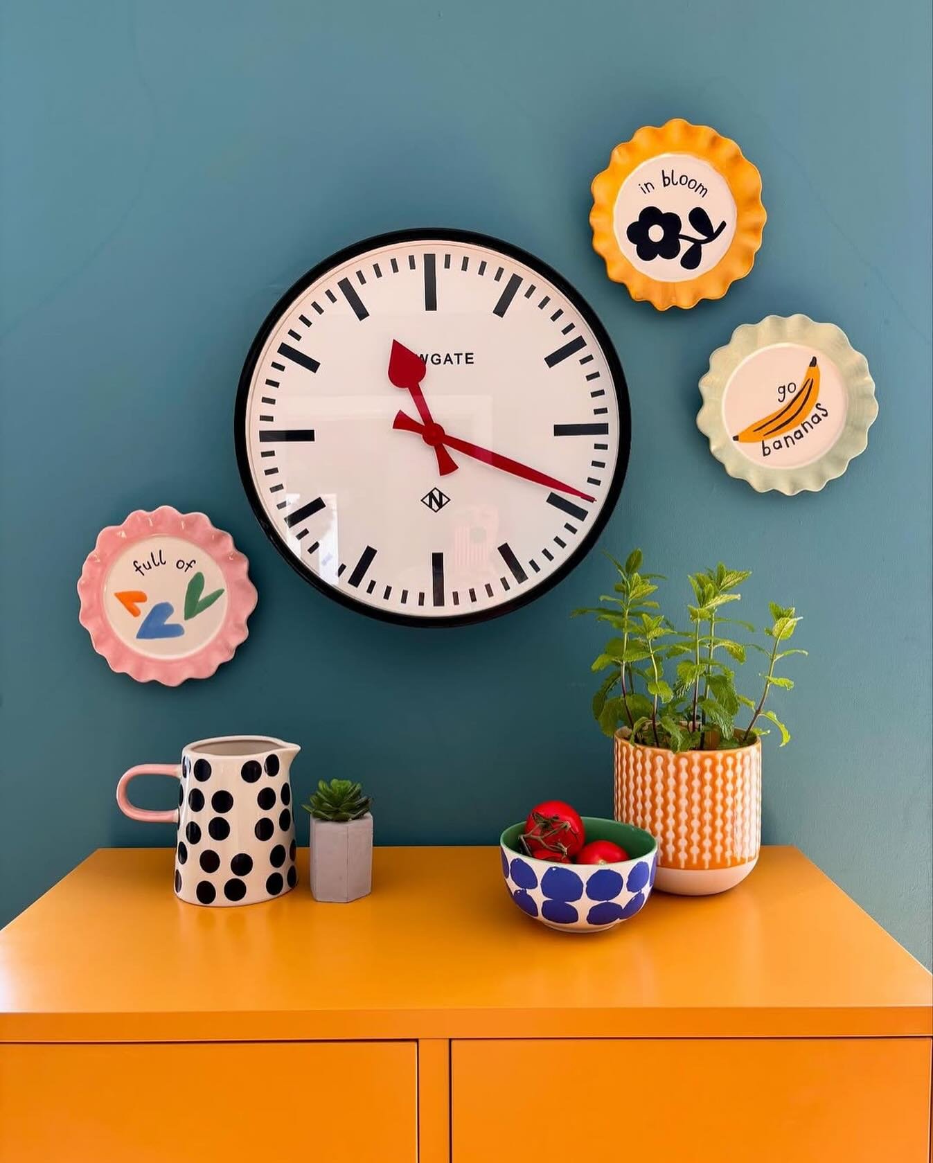 Teal wall featuring a large white Newgate clock surrounded by small colorful whimsical plates above an orange cabinet