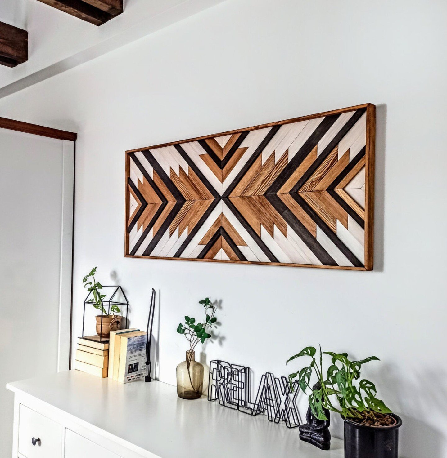 Rectangular wooden wall art with black white and brown chevron patterns hanging above a white dresser with plants
