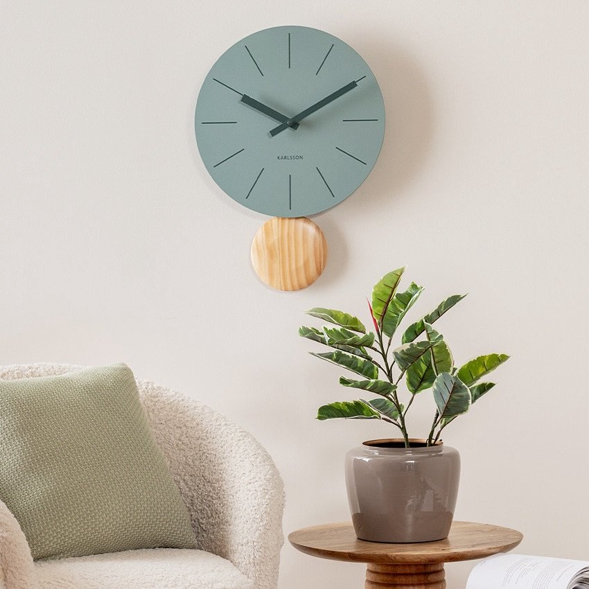 Sage green wall clock with wooden pendulum on a white wall above a beige armchair