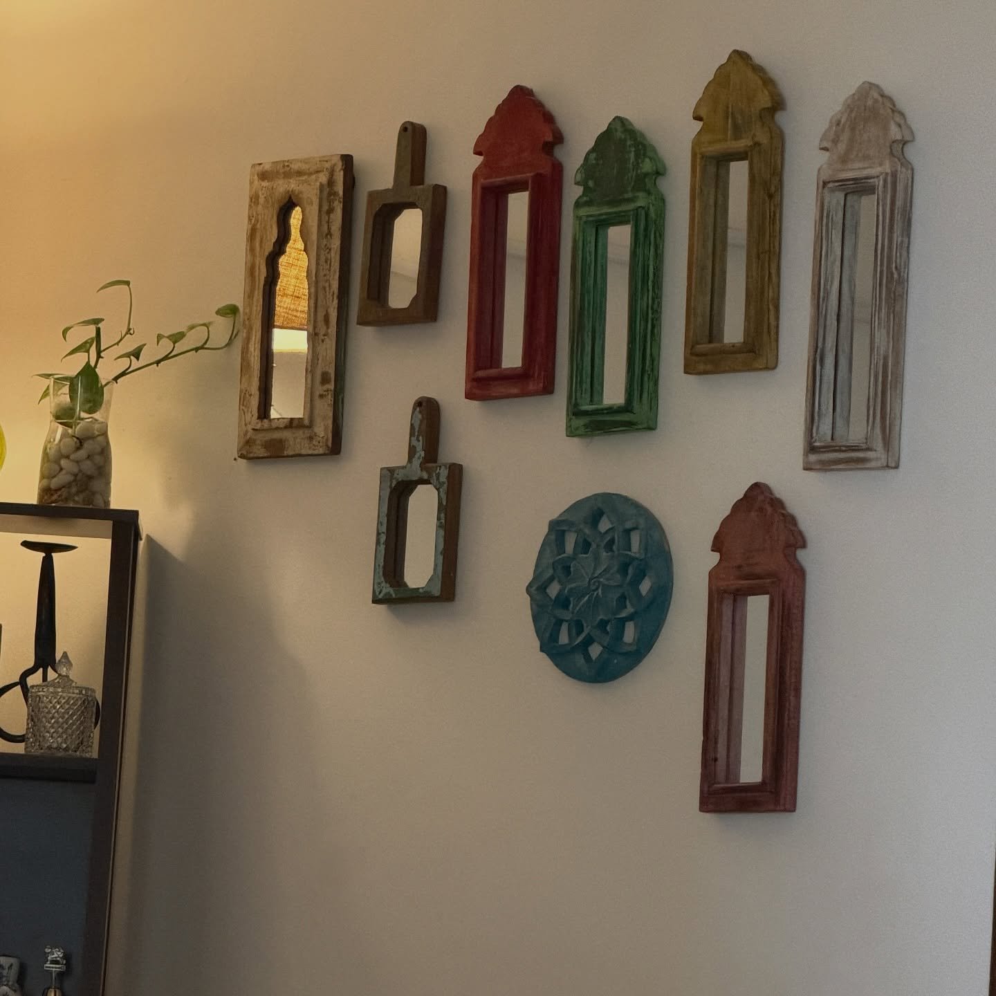 Wall display of small colorful distressed wooden mirrors shaped like windows arranged on a white wall next to a shelf.