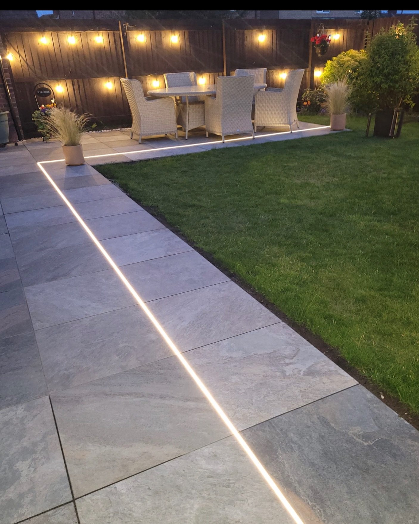 Backyard garden patio featuring a recessed linear LED strip separating the grey paving stones from the grass law.