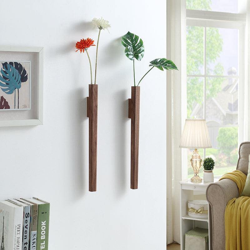 Two slender vertical wooden wall vases holding flower stems on a white wall near books