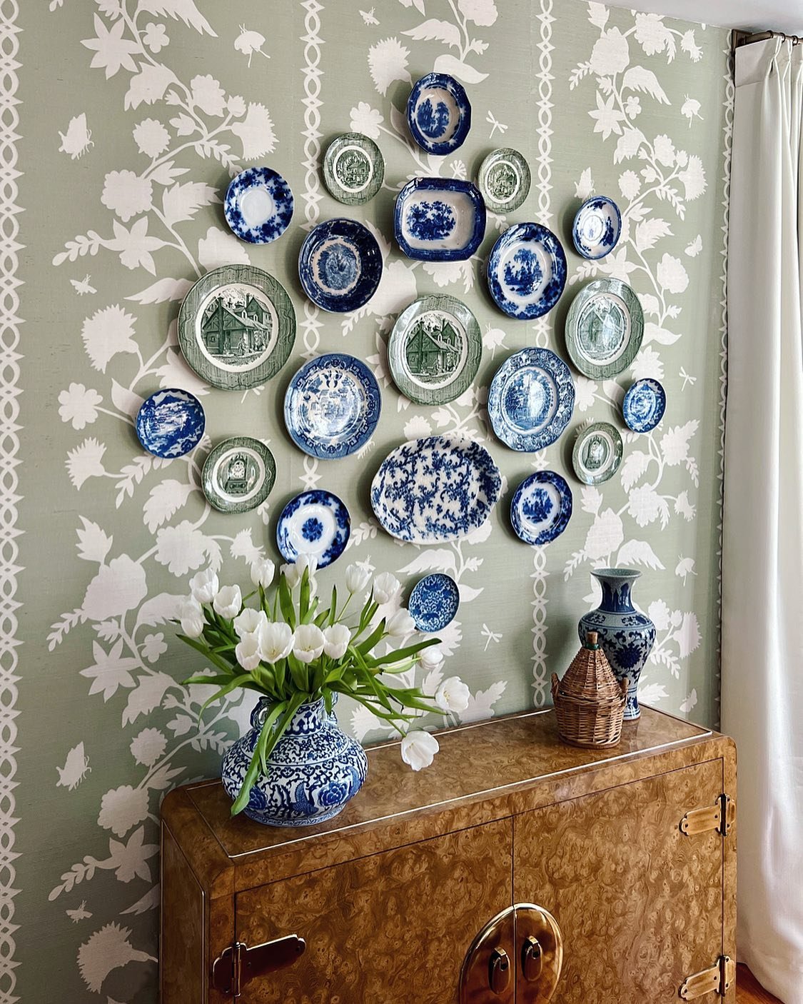 Green floral wallpaper wall featuring a mixed collection of blue and green plates above a burl wood console