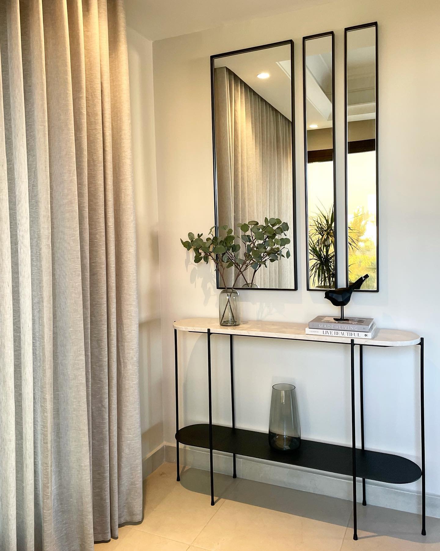 Three vertical rectangular mirrors with black frames hanging above a marble console table next to beige curtains.