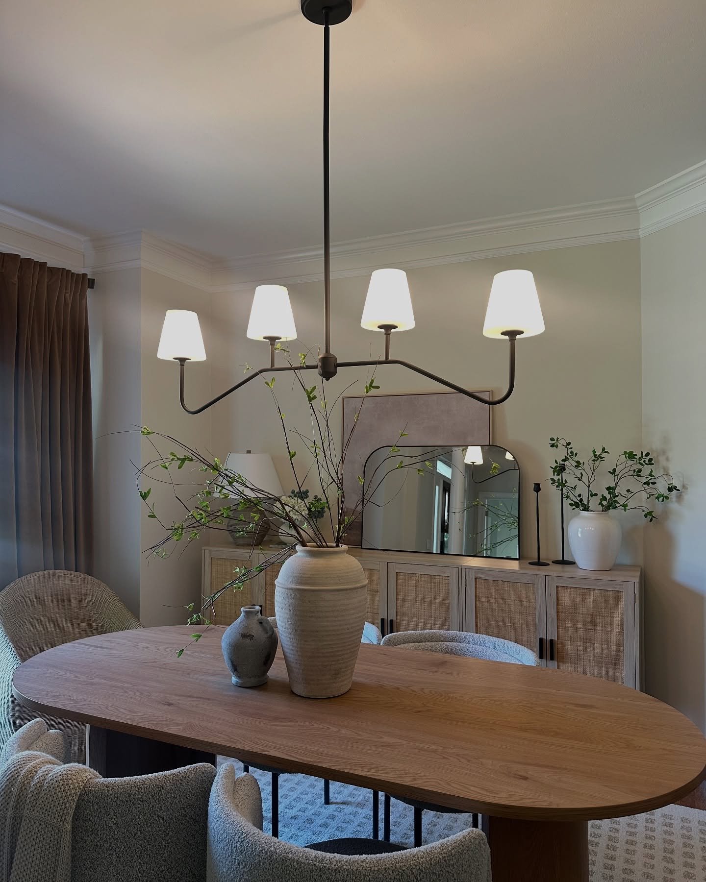 Black curved arm chandelier with white shades hanging over an oval wood dining table with green branches.