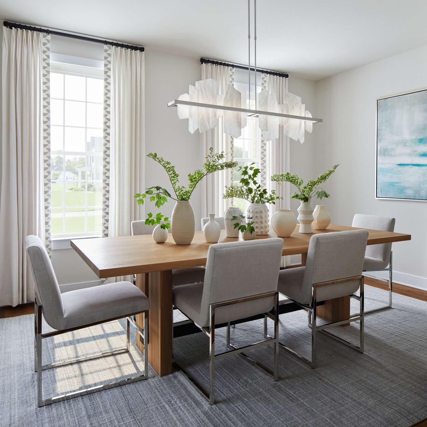 Modern linear alabaster chandelier hanging over a light wood dining table with grey chairs.