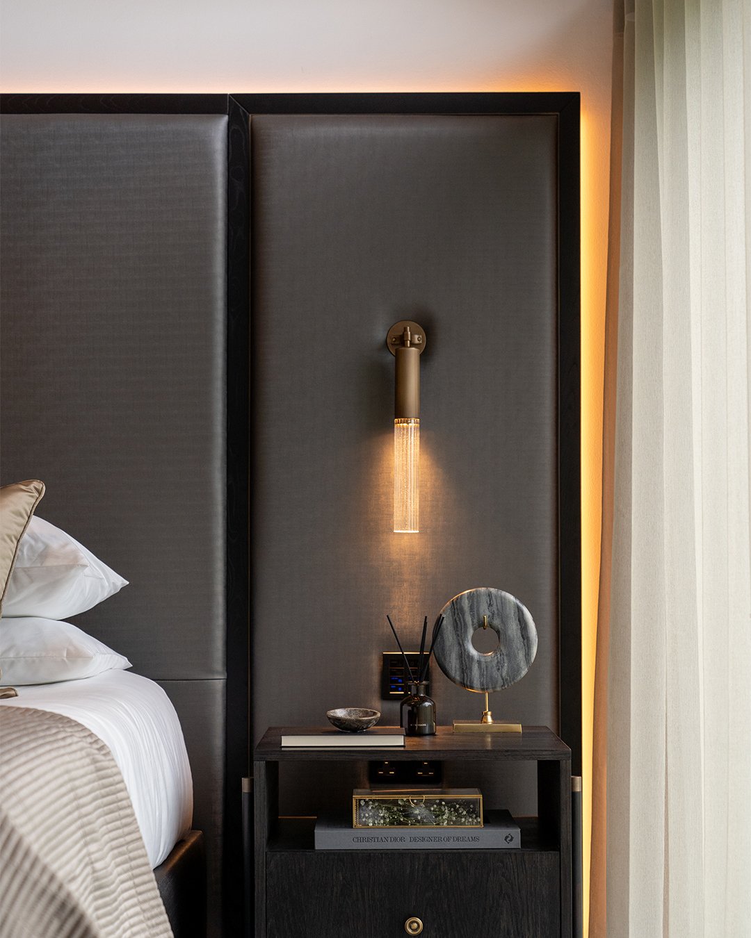 Bedroom accent wall featuring a dark upholstered headboard with warm LED backlighting and a brass sconce.