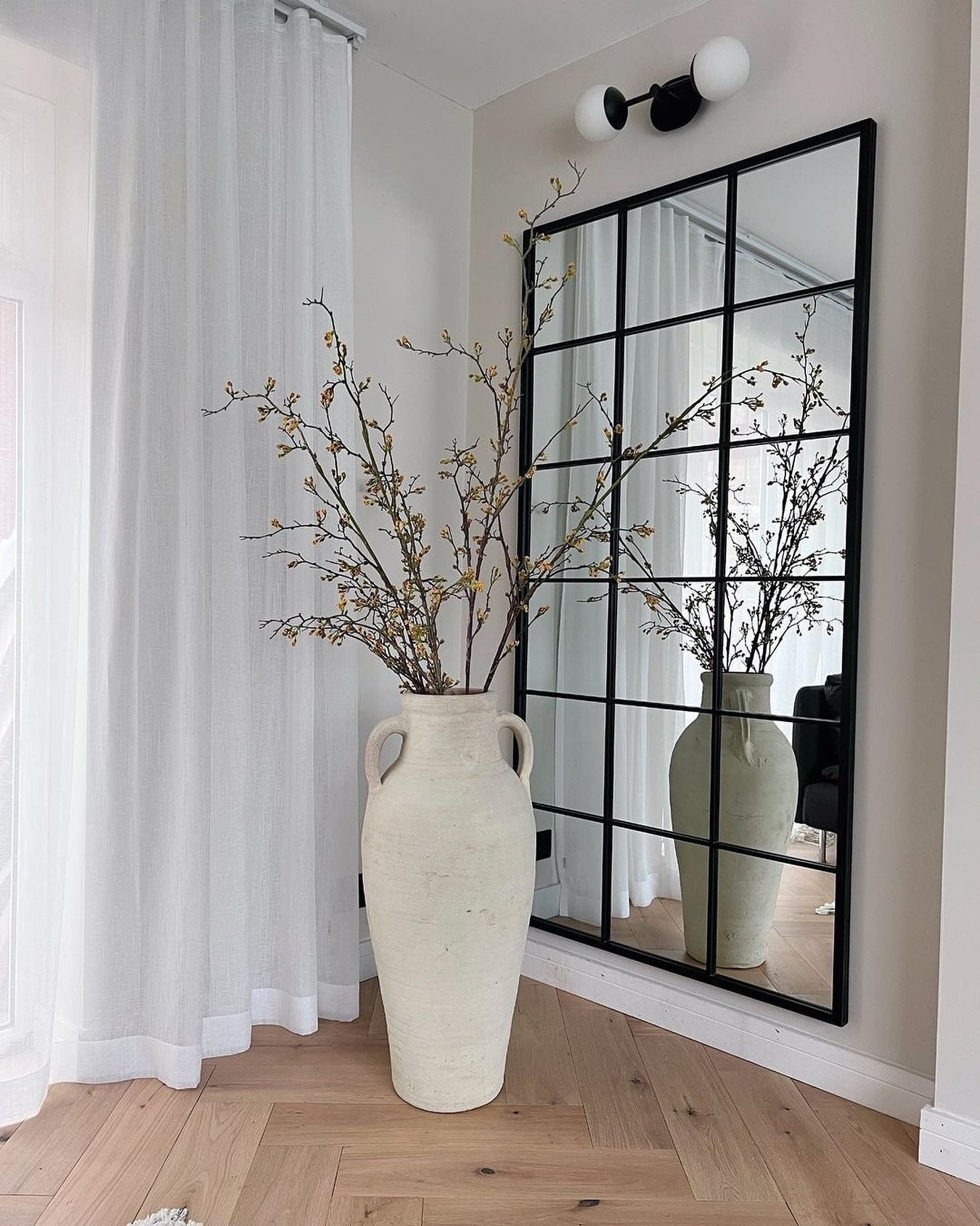 Large black windowpane mirror leaning against a corner wall behind an oversized rustic beige vase with tall branches.