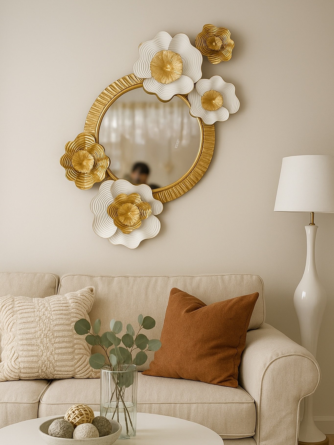 Round gold mirror with white floral frame accents hanging above a beige sofa with rust-colored pillows.