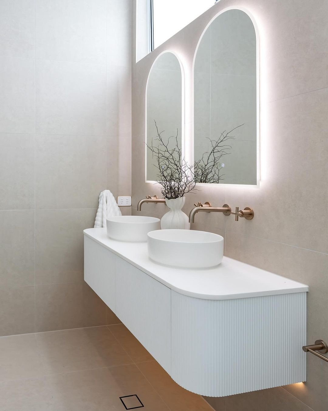 Beige tiled bathroom wall with two backlit arched mirrors above white fluted vanity