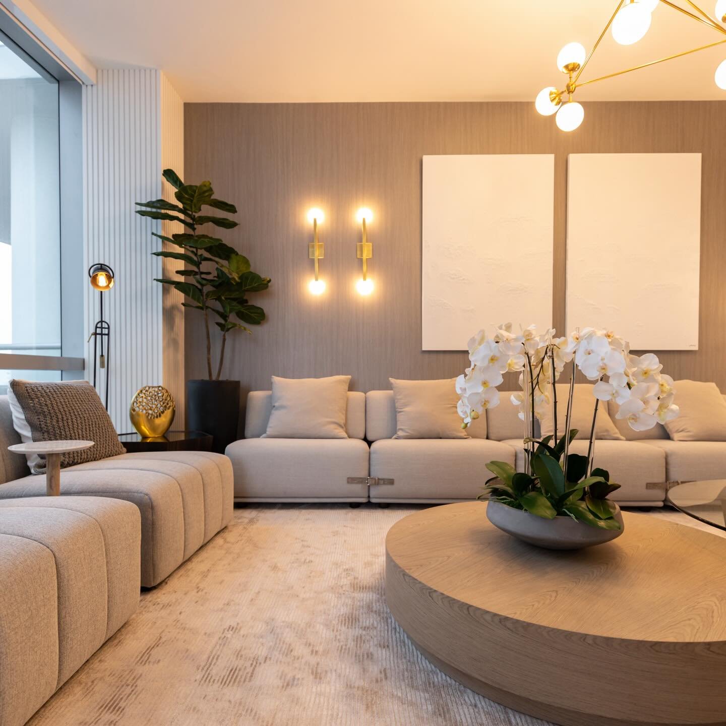 Modern living room with a grey textured accent wall featuring two white canvases and a gold double-bulb wall sconce behind a beige sofa.