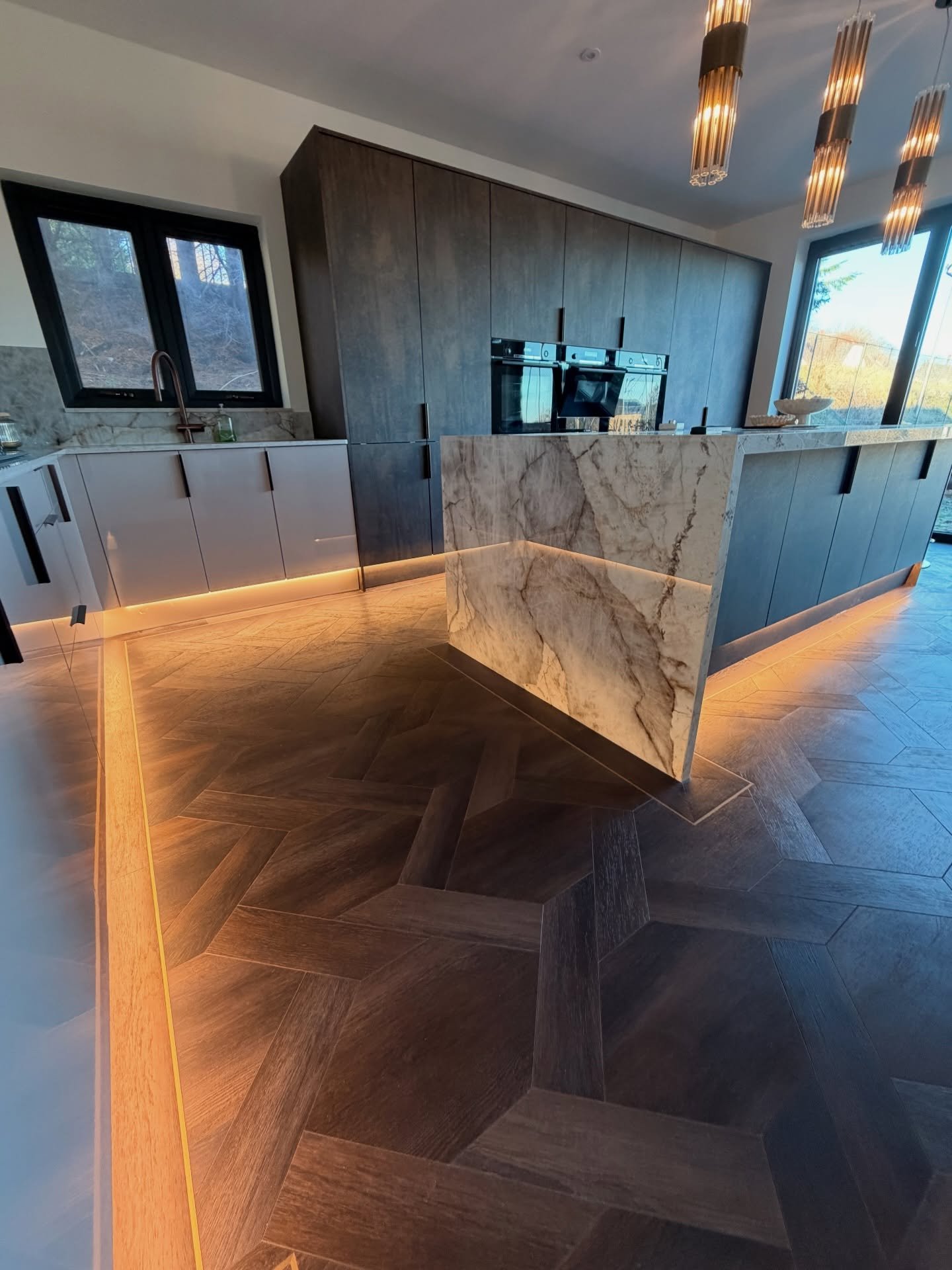 Modern kitchen with warm LED strips recessed in herringbone flooring and under the marble island toe kick.